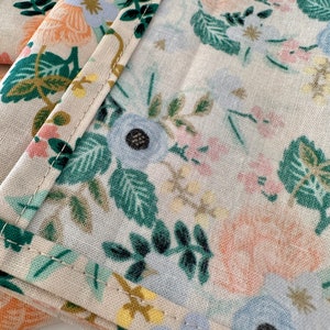 May include: Close-up of a beige fabric with a floral pattern. The fabric features a variety of flowers in shades of blue, pink, and orange, as well as green leaves. The fabric is sewn with a white thread.