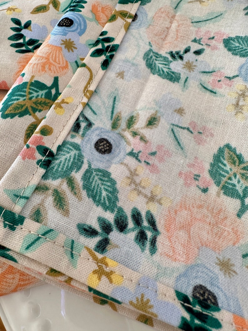 May include: Close-up of a beige fabric with a floral pattern. The fabric features a variety of flowers in shades of blue, pink, and orange, as well as green leaves. The fabric is sewn with a white thread.