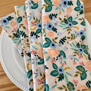 May include: Four folded fabric napkins with a floral pattern. The napkins are beige with a repeating pattern of blue, orange, green, and pink flowers.