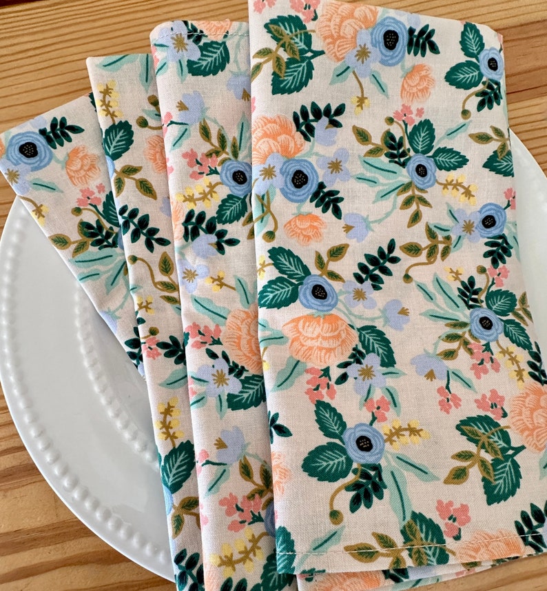 May include: Four folded fabric napkins with a floral pattern. The napkins are beige with a repeating pattern of blue, orange, green, and pink flowers.