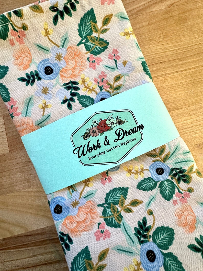May include: A set of white cotton napkins with a floral pattern in shades of blue, pink, and yellow. The napkins are packaged with a blue and white label that reads "Work & Dream Everyday Cotton Napkins".