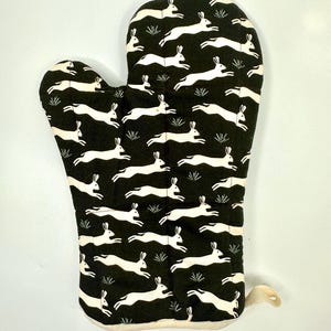 May include: Black oven mitt with a repeating pattern of white running hares and small green plants. The mitt has a cream-colored trim and a loop for hanging. A kitchen accessory.