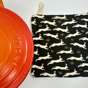 May include: A black and white quilted pot holder featuring a repeating pattern of running rabbits. The pot holder has a cream-colored loop for hanging. An orange Le Creuset pot is partially visible in the image.