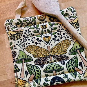 Puede incluir: Un posavasos de color crema con un patr&oacute;n caprichoso de setas verdes, mariposas amarillas y flores amarillas. El posavasos tiene un lazo para colgar.