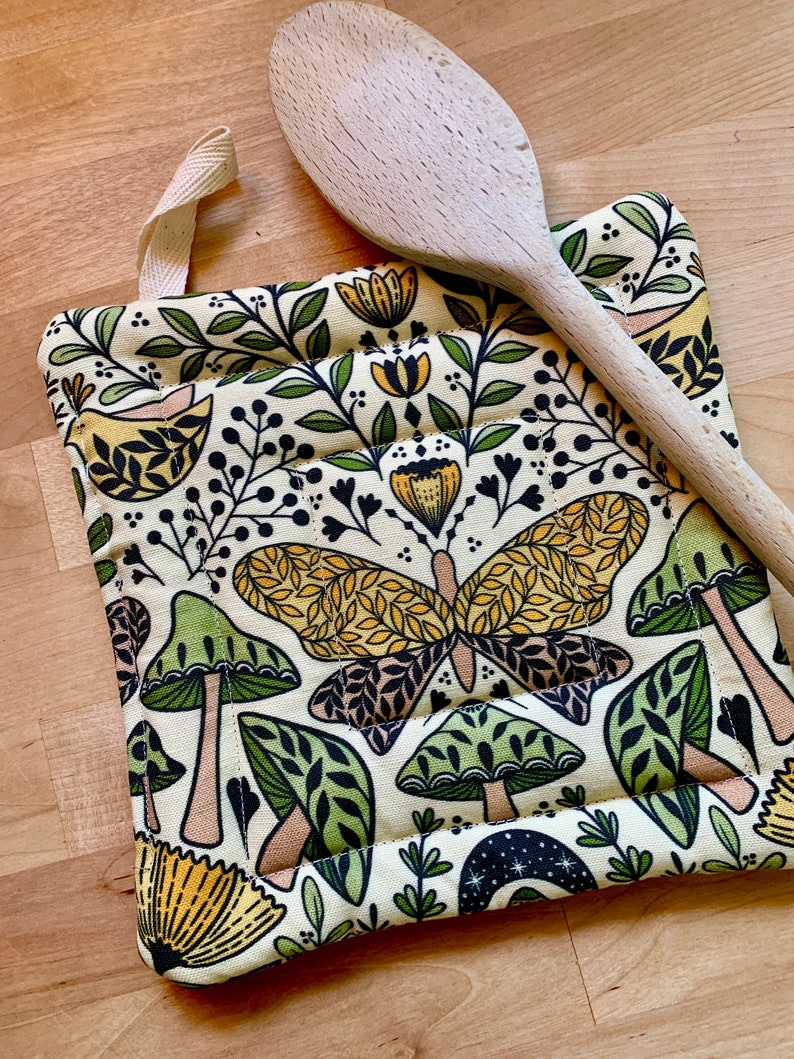 Puede incluir: Un posavasos de color crema con un patr&oacute;n caprichoso de setas verdes, mariposas amarillas y flores amarillas. El posavasos tiene un lazo para colgar.