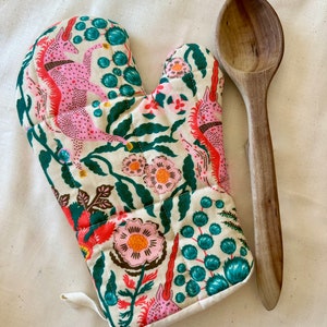 May include: A white oven mitt with a floral pattern featuring pink unicorns, green leaves, and colorful flowers. The mitt has a loop for hanging and a wooden spoon is lying next to it.