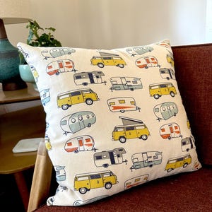 May include: A white throw pillow with a pattern of yellow and blue vintage camper vans and trailers. The pillow is on a red couch.