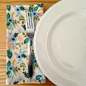 May include: A white ceramic dinner plate with a beaded edge sits on a wooden table next to a floral patterned napkin and a silver fork.