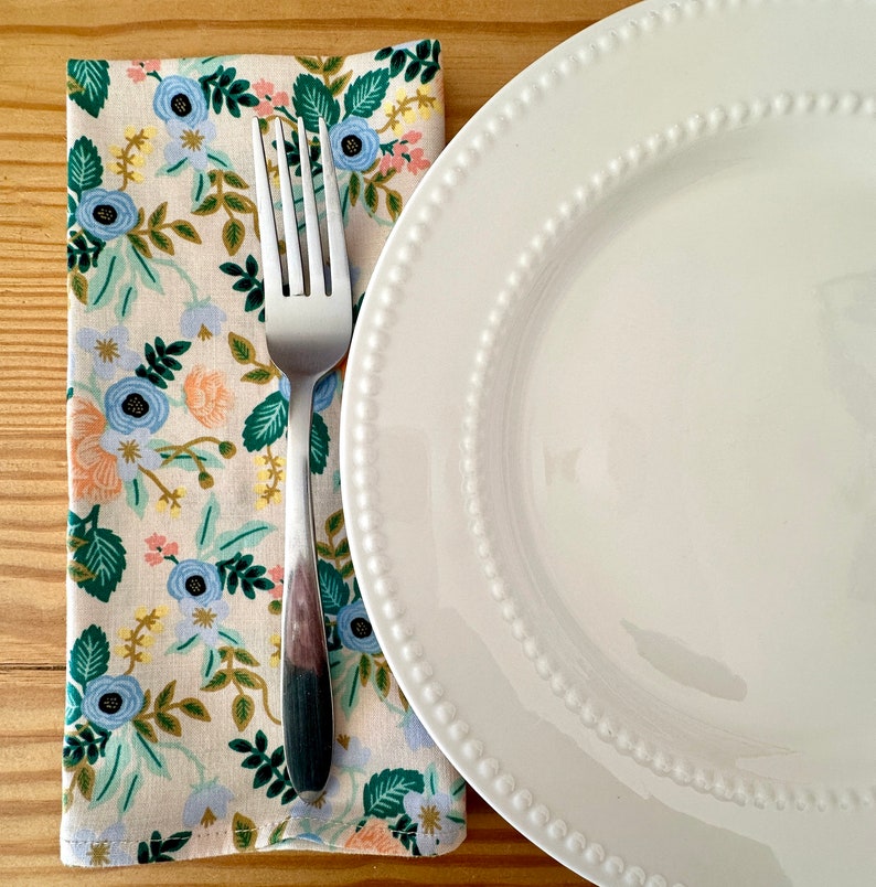 May include: A white ceramic dinner plate with a beaded edge sits on a wooden table next to a floral patterned napkin and a silver fork.