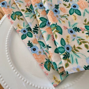 May include: Four folded fabric napkins with a floral pattern. The napkins are white with a light blue, peach, and green floral design.