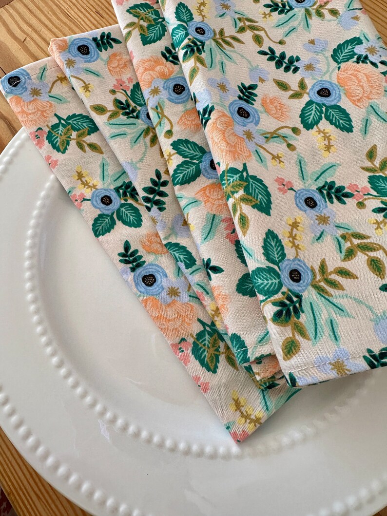 May include: Four folded fabric napkins with a floral pattern. The napkins are white with a light blue, peach, and green floral design.