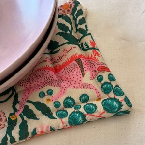May include: A pink plate rests on a patterned trivet. The trivet features a cream background with a pink and orange mythical creature design, along with green foliage and teal accents. The trivet is quilted and has a stitched edge.