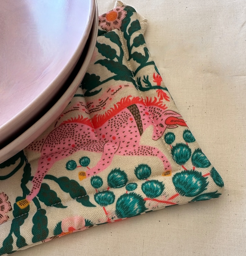 May include: A pink plate rests on a patterned trivet. The trivet features a cream background with a pink and orange mythical creature design, along with green foliage and teal accents. The trivet is quilted and has a stitched edge.