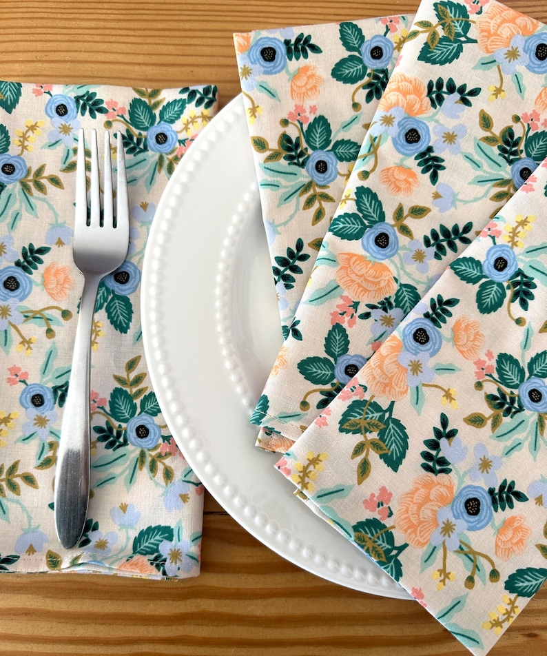 May include: Three floral patterned napkins in shades of blue, green, peach, and yellow on a white plate with a silver fork.