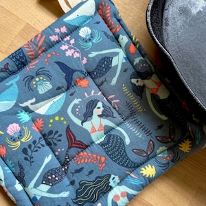 May include: A blue quilted pot holder with a mermaid and sea life pattern. The pot holder has a loop for hanging.