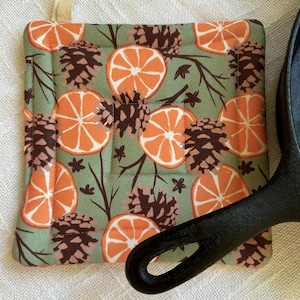 May include: A green fabric pot holder with a pattern of orange slices and pine cones. The pot holder is quilted and has a brown binding. A black cast iron skillet is resting on the pot holder.