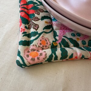 May include: A floral patterned pot holder with pink, orange, and green flowers on a cream background. Two pink plates rest on the pot holder. The pot holder has a quilted design.