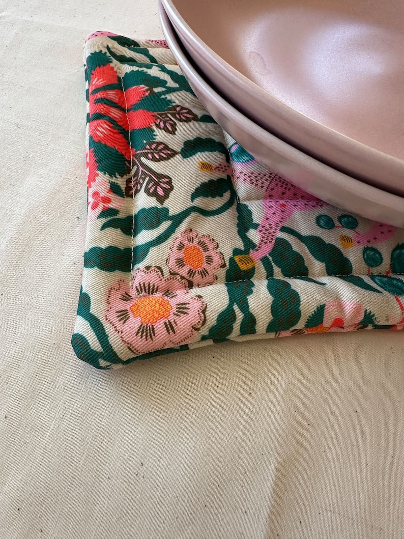 May include: A floral patterned pot holder with pink, orange, and green flowers on a cream background. Two pink plates rest on the pot holder. The pot holder has a quilted design.