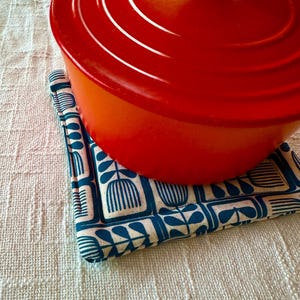 May include: A vibrant red enameled cast iron pot rests on a square, patterned trivet. The trivet features a repeating design of blue and white stylized floral shapes. The pot has a rounded shape with a lid and side handles.
