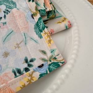 May include: A set of folded fabric napkins with a floral pattern in shades of blue, green, yellow, and pink. The napkins have a white background and are folded in a triangular shape.