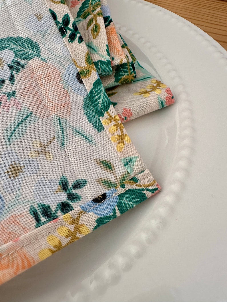 May include: A set of folded fabric napkins with a floral pattern in shades of blue, green, yellow, and pink. The napkins have a white background and are folded in a triangular shape.