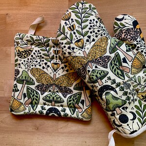 Puede incluir: Un par de guantes para horno beige con un dise&ntilde;o floral y de mariposa. Los guantes est&aacute;n hechos de tela acolchada y tienen un lazo para colgar.