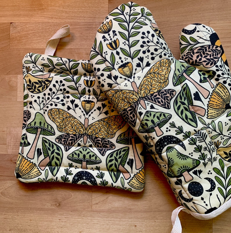 Puede incluir: Un par de guantes para horno beige con un dise&ntilde;o floral y de mariposa. Los guantes est&aacute;n hechos de tela acolchada y tienen un lazo para colgar.