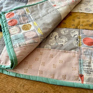 May include: A patchwork quilt with a variety of patterns and colors, including pink, gray, and yellow. The quilt is folded in half, revealing the back side, which is a solid pink with a geometric pattern.