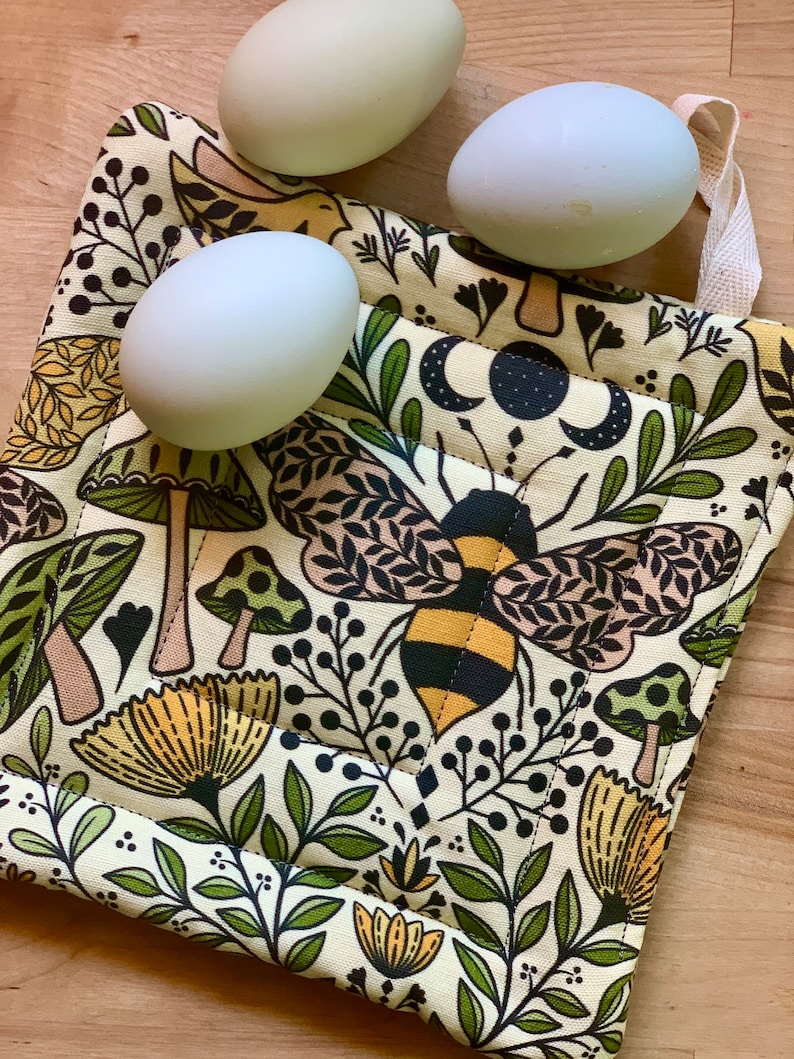 Puede incluir: Un agarrador de olla de tela beige con una abeja, setas y dise&ntilde;os florales. El agarrador de olla tiene un asa de bucle blanco.