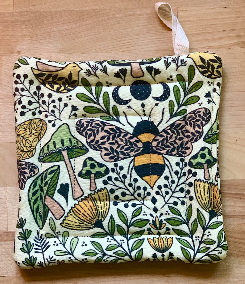 Puede incluir: Un agarrador de olla acolchado verde, amarillo y marr&oacute;n con una abeja, setas y dise&ntilde;os florales.