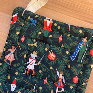 May include: A square, quilted pot holder with a dark green background featuring a festive Nutcracker-themed pattern. The design includes illustrations of toy soldiers, ballerinas, and Christmas ornaments. A cream-colored loop is attached for hanging.