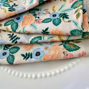 May include: A stack of three cream-colored tea towels with a floral pattern. The pattern features blue, green, orange, and pink flowers on a cream background.