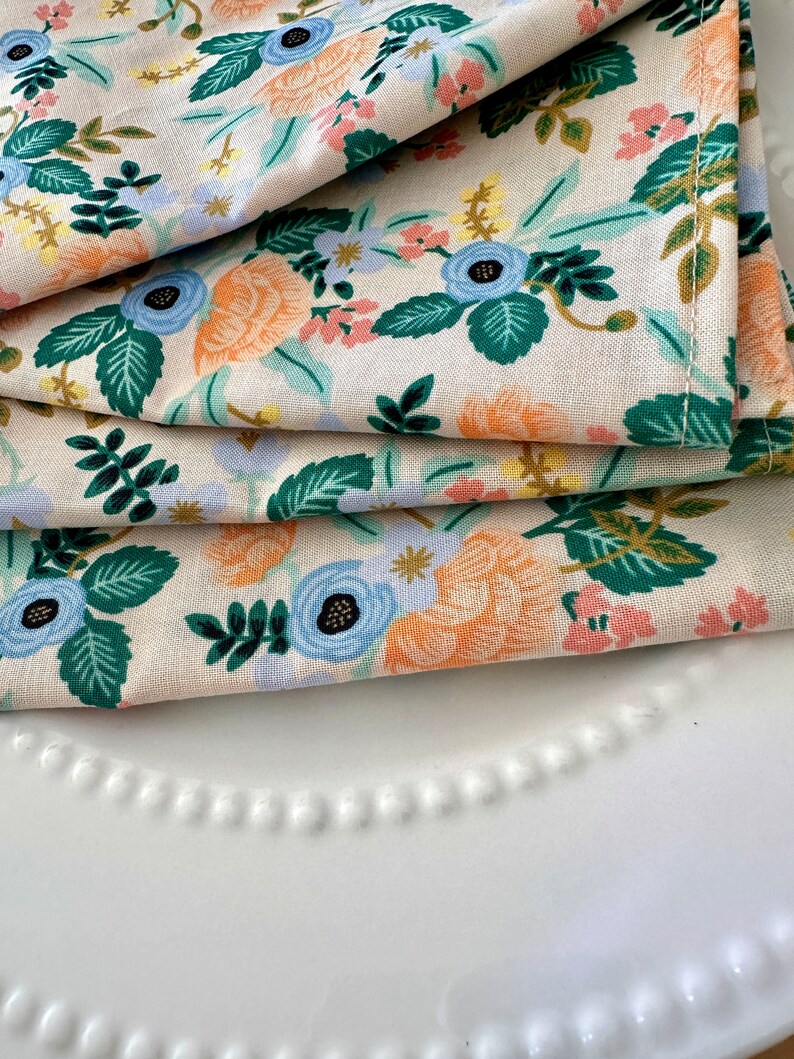 May include: A stack of three cream-colored tea towels with a floral pattern. The pattern features blue, green, orange, and pink flowers on a cream background.