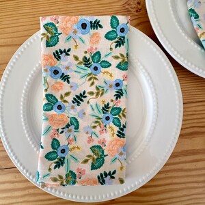 May include: A white dinner plate with a beaded edge and a floral patterned napkin folded in the center. The napkin has a light pink background with blue, green, and orange flowers.