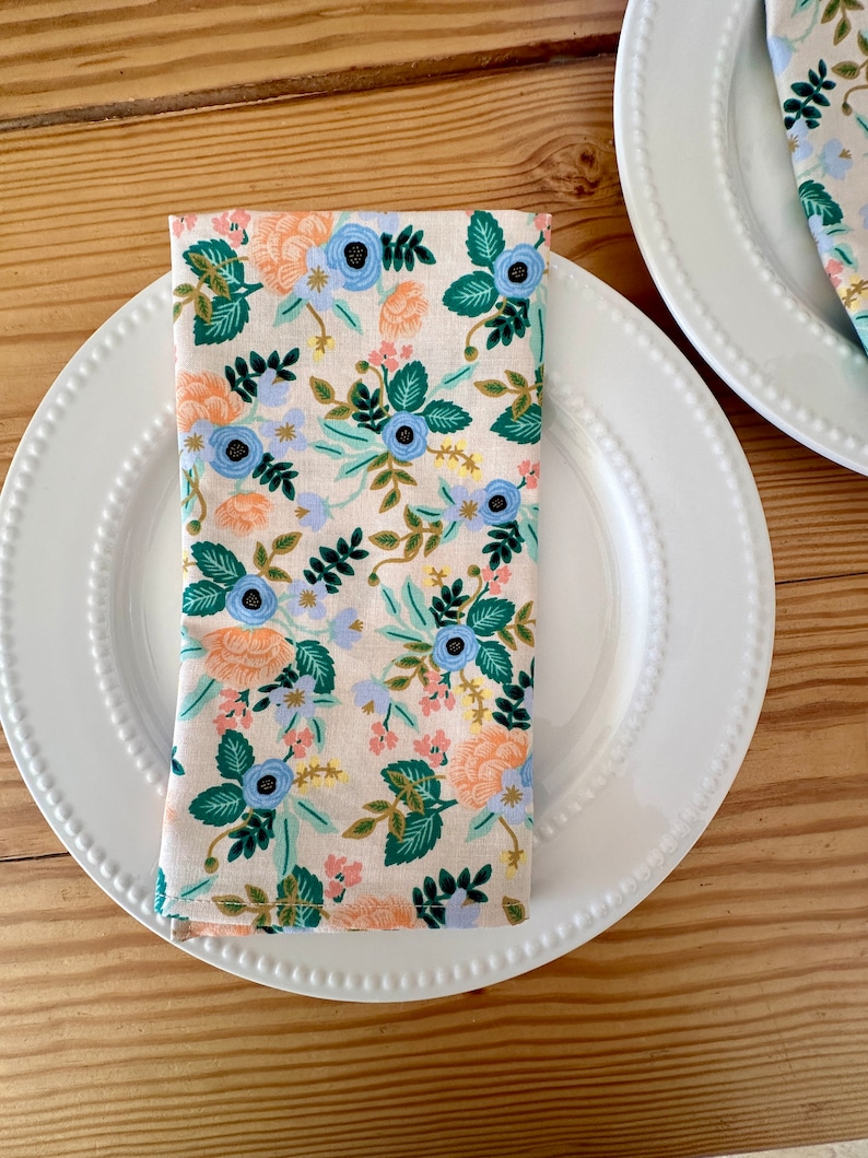 May include: A white dinner plate with a beaded edge and a floral patterned napkin folded in the center. The napkin has a light pink background with blue, green, and orange flowers.