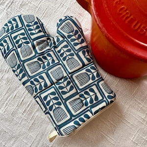May include: A teal and white patterned oven mitt with a loop for hanging. The pattern features stylized floral and leaf designs. A red cooking pot is partially visible in the background, suggesting a kitchen theme.