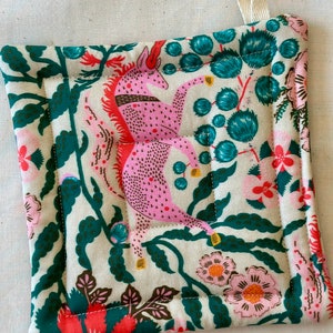 May include: Square quilted pot holder with a cream background featuring pink unicorns, teal floral accents, and pink and orange flowers. The edges are trimmed with a dark green fabric. A cream-colored loop is attached for hanging.