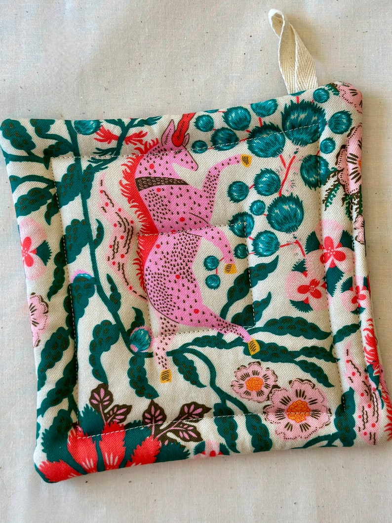 May include: Square quilted pot holder with a cream background featuring pink unicorns, teal floral accents, and pink and orange flowers. The edges are trimmed with a dark green fabric. A cream-colored loop is attached for hanging.