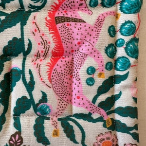 May include: Close-up of a quilted fabric with a repeating pattern. The design features a pink mythical creature with red accents, surrounded by dark green foliage and pink flowers on a cream background. The edges are neatly stitched.