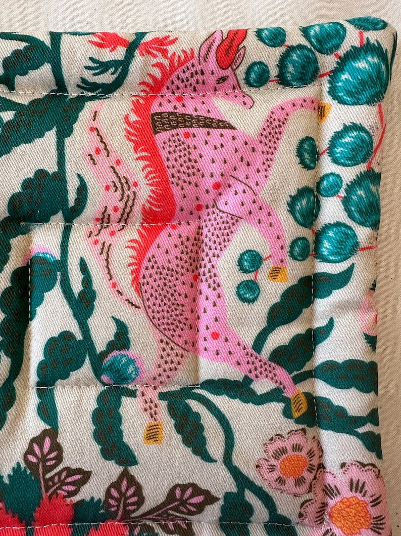 May include: Close-up of a quilted fabric with a repeating pattern. The design features a pink mythical creature with red accents, surrounded by dark green foliage and pink flowers on a cream background. The edges are neatly stitched.