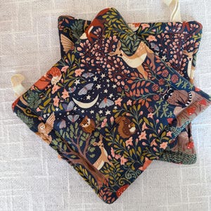 May include: Two square quilted pot holders featuring a dark blue background with a forest-themed print. The pattern includes illustrations of deer, owls, squirrels, moths, crescent moons, stars, mushrooms, and various green foliage with pink flowers.