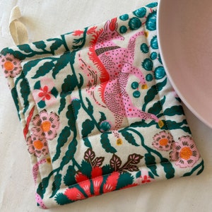 May include: Square, quilted pot holder with a cream background and a colorful floral and mythical creature design. The design includes pink, coral, teal, and dark green elements. A cream-colored loop is attached for hanging.