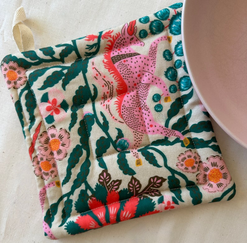 May include: Square, quilted pot holder with a cream background and a colorful floral and mythical creature design. The design includes pink, coral, teal, and dark green elements. A cream-colored loop is attached for hanging.