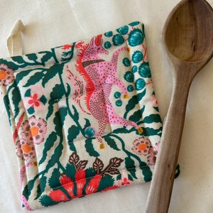 May include: Quilted pot holder with a pink mythical creature design, surrounded by floral and leaf patterns in shades of green, red, and cream. A wooden spoon rests beside the pot holder. The pot holder has a loop for hanging.