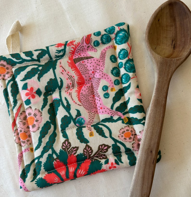 May include: Quilted pot holder with a pink mythical creature design, surrounded by floral and leaf patterns in shades of green, red, and cream. A wooden spoon rests beside the pot holder. The pot holder has a loop for hanging.