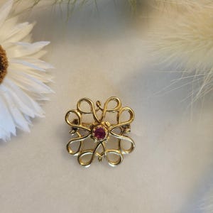 Puede incluir: Un broche de tono dorado con un diseño delicado e intrincado que presenta una piedra preciosa rosa central.