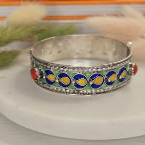 May include: Silver-tone hinged bracelet with a decorative enamel design. The bracelet features a green band with blue and yellow accents, and red gemstone details. A stylish piece of jewelry.
