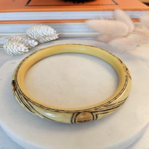 May include: Cream-coloured bangle bracelet with dark brown etched designs. The bracelet has a rounded shape and is made of a smooth material. The etched designs include geometric and curved lines. The bracelet is displayed on a white surface.