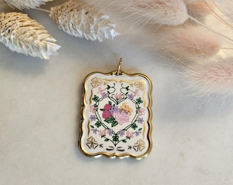 May include: A gold-toned pendant with a white porcelain insert featuring a floral design. The design includes a heart-shaped arrangement of pink, purple, and yellow flowers surrounded by green leaves and gold accents.