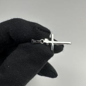 Może przedstawiać: Srebrny wisiorek w kształcie krzyża z małą pętelką na łańcuszek. Krzyż ma prosty, minimalistyczny design z poziomym paskiem przecinającym pasek pionowy. Wisiorek jest oznaczony napisem "925".
